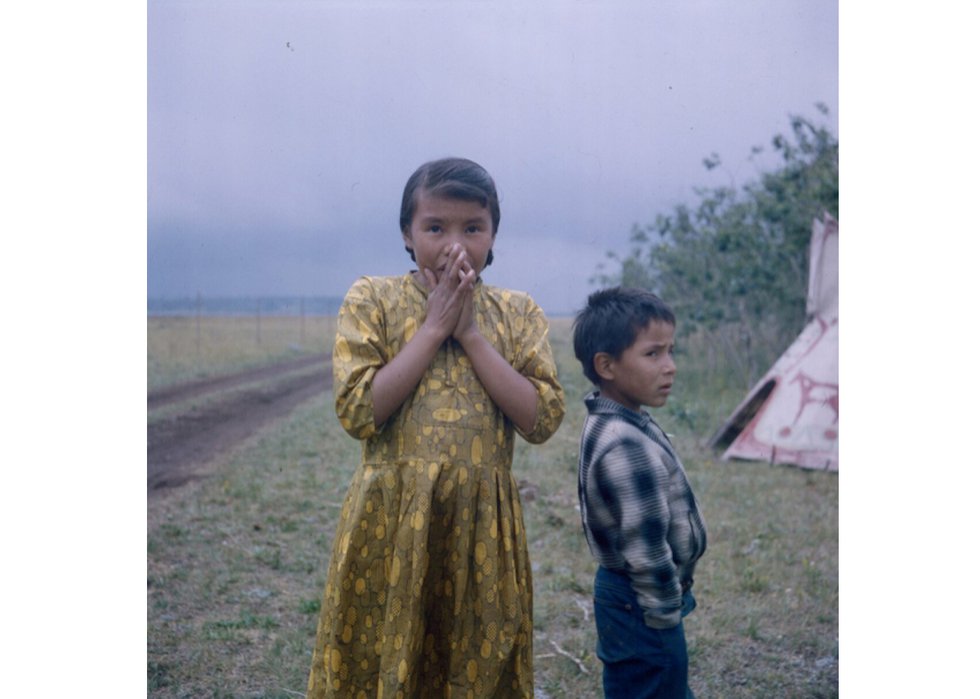 Young girl and boy standing on grass, between a teepee and a dirt road, Ghost River, Alberta, ca. 1962